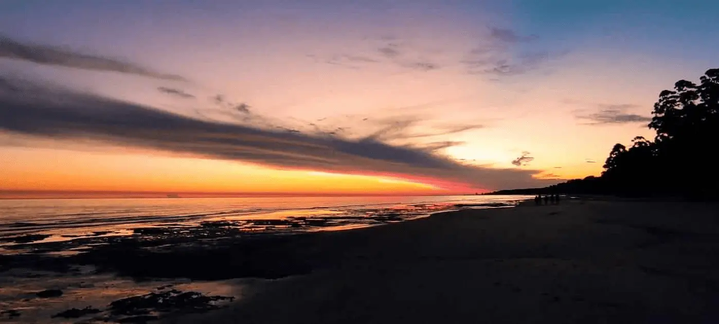 A panoramic view of a beach at sunset, showcasing a breathtaking horizon where a vibrant orange and yellow sky meets a calm sea. A dramatic cloud formation stretches across the sky, casting shadows and creating a contrast with the glowing sunset. The beach is lined with dark silhouettes of trees on the right, and there are small figures of people enjoying the serene evening. The scene captures a peaceful and majestic end to the day, highlighted by the natural beauty of the shoreline and the spectacular sky.