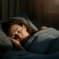A woman with wavy brown hair is peacefully sleeping in a dimly lit bedroom. She is lying on her side with her head resting on a soft pillow, her face relaxed, and a slight smile on her lips. A cozy gray blanket covers her, adding to the warmth and comfort of the scene. The room has a calm ambiance with soft lighting, possibly from a nearby window or a bedside lamp, casting gentle shadows. The overall mood conveys rest, relaxation, and tranquility.