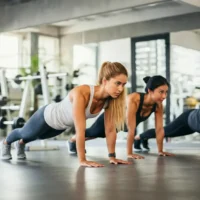 Two fit women are performing push-ups in a well-equipped gym. The woman in the foreground has long blonde hair tied in a ponytail and is wearing a white tank top and dark leggings. She has a focused and determined expression as she maintains a strong plank position. Behind her, another woman with dark hair tied in a bun, wearing a black sports bra and dark leggings, is also engaged in the same exercise. The gym has large windows allowing natural light to enter, with various workout equipment visible in the background, creating a motivating and energetic fitness atmosphere.