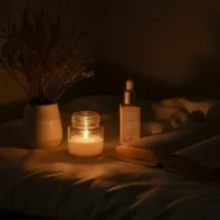 A cozy nighttime scene featuring a softly lit candle on a bed, casting a warm glow. Beside the candle, there is an open book with its pages slightly curled, a bottle of skincare serum with a dropper, and a white ceramic vase holding dried flowers. The background is dimly lit with plush bedding visible, creating a peaceful and relaxing ambiance. The composition evokes a sense of calm, self-care, and unwinding before bedtime.