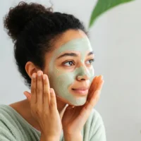 A young woman with curly dark hair tied up in a bun is applying a green facial mask to her face. She is gently touching her cheeks with both hands and has a relaxed, content expression. She is wearing a soft, light green bathrobe, and the background features a blurred indoor setting with a large green plant leaf visible. The lighting is soft and natural, enhancing the fresh and self-care ambiance of the image.