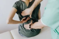 A physical therapist or chiropractor in a green medical uniform is examining a patient's back. The patient, a woman wearing a dark gray t-shirt, is seated on an examination table and pointing to a specific area on her lower back while the therapist places their hands on her back to assess the issue. The image conveys a medical or therapeutic setting focused on back pain or spinal health.