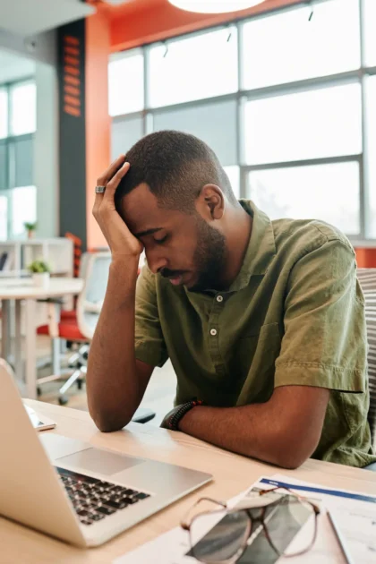 A professional man in his 30s feeling tired, stressed, depleated with his hand on his forehead at his desk in front of his laptop