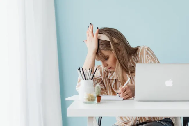 A young blond professional woman in her 30s placing her hand on her forehead as she feels stuck, frustrated, tired in front of her laptop at her desk